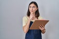 Hispanic girl wearing apron holding clipboard serious face thinking about question with hand on chin, thoughtful about confusing