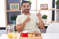 Hispanic man with beard eating breakfast smiling and looking at the camera pointing with two hands and fingers to the side