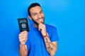 Hispanic man with beard holding italy passport with hand on chin thinking about question, pensive expression