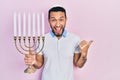 Hispanic man with beard holding menorah hanukkah jewish candle pointing thumb up to the side smiling happy with open mouth Royalty Free Stock Photo