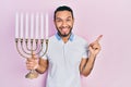 Hispanic man with beard holding menorah hanukkah jewish candle smiling happy pointing with hand and finger to the side Royalty Free Stock Photo
