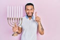 Hispanic man with beard holding menorah hanukkah jewish candle smiling with an idea or question pointing finger with happy face, Royalty Free Stock Photo