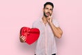 Hispanic man with beard holding valentine gift serious face thinking about question with hand on chin, thoughtful about confusing