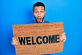 Hispanic man with beard holding welcome doormat afraid and shocked with surprise and amazed expression, fear and excited face Royalty Free Stock Photo