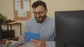 Hispanic man with beard in an office holding a tablet, surrounded by computer, documents, plants, and office supplies, conveying a Royalty Free Stock Photo