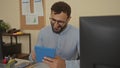 Hispanic man with beard in an office holding a tablet, surrounded by computer, documents, plants, and office supplies, conveying a Royalty Free Stock Photo