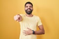 Hispanic man with beard standing over yellow background laughing at you, pointing finger to the camera with hand over body, shame Royalty Free Stock Photo