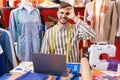 Hispanic man with beard using laptop at tailor room smiling happy doing ok sign with hand on eye looking through fingers