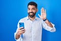 Hispanic man with beard using smartphone typing message waiving saying hello happy and smiling, friendly welcome gesture Royalty Free Stock Photo