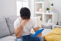 Hispanic man with beard using touchpad sitting on the sofa covering eyes with hand, looking serious and sad Royalty Free Stock Photo