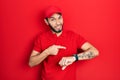 Hispanic man with beard wearing delivery uniform and cap in hurry pointing to watch time, impatience, upset and angry for deadline