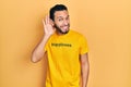 Hispanic man with beard wearing t shirt with happiness word message smiling with hand over ear listening an hearing to rumor or