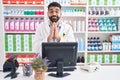 Hispanic man with beard working at pharmacy drugstore praying with hands together asking for forgiveness smiling confident