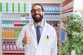 Hispanic man with beard working at pharmacy drugstore smiling happy and positive, thumb up doing excellent and approval sign