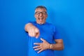 Hispanic man with grey hair standing over blue background laughing at you, pointing finger to the camera with hand over body, Royalty Free Stock Photo