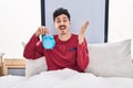 Hispanic man holding alarm clock in the bed celebrating victory with happy smile and winner expression with raised hands Royalty Free Stock Photo