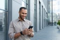 hispanic man outside modern office building using smartphone, businessman in shirt typing message and browsing online Royalty Free Stock Photo