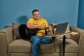 Hispanic man on a sofa in front of his laptop Greeting fellow students in the online guitar class
