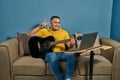 Hispanic man on a sofa in front of his laptop Greeting fellow students in the online guitar class