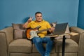 Hispanic man on a sofa in front of his laptop Greeting fellow students in the online guitar class