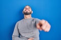 Hispanic man standing over blue background laughing at you, pointing finger to the camera with hand over body, shame expression Royalty Free Stock Photo