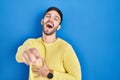 Hispanic man standing over blue background laughing at you, pointing finger to the camera with hand over body, shame expression Royalty Free Stock Photo