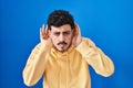 Hispanic man standing over blue background trying to hear both hands on ear gesture, curious for gossip Royalty Free Stock Photo