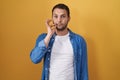 Hispanic man standing over yellow background mouth and lips shut as zip with fingers