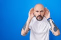 Hispanic man with tattoos standing over blue background trying to hear both hands on ear gesture, curious for gossip Royalty Free Stock Photo