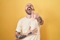 Hispanic man with tattoos standing over yellow background laughing at you, pointing finger to the camera with hand over body, Royalty Free Stock Photo
