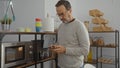 Hispanic man using smartphone in a cozy kitchenette setting with modern decor, surrounded by shelves, jars, and utensils, creating Royalty Free Stock Photo