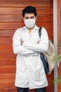 Hispanic medical student wearing a facemask and a medical robe and standing against a wooden wall