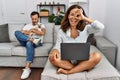 Hispanic middle age couple at home, woman using laptop doing ok gesture with hand smiling, eye looking through fingers with happy Royalty Free Stock Photo