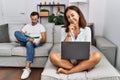 Hispanic middle age couple at home, woman using laptop looking confident at the camera with smile with crossed arms and hand Royalty Free Stock Photo