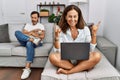 Hispanic middle age couple at home, woman using laptop shouting with crazy expression doing rock symbol with hands up Royalty Free Stock Photo