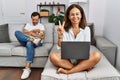 Hispanic middle age couple at home, woman using laptop smiling with happy face winking at the camera doing victory sign Royalty Free Stock Photo