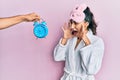 Hispanic teenager girl with dental braces wearing sleep mask and robe with alarm clock close to her shouting and screaming loud to Royalty Free Stock Photo