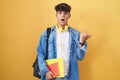 Hispanic teenager wearing student backpack and holding books surprised pointing with hand finger to the side, open mouth amazed Royalty Free Stock Photo