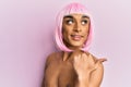 Hispanic transgender man wearing make up and pink wig smiling with happy face looking and pointing to the side with thumb up