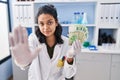 Hispanic woman with dark hair working at scientist laboratory holding money with open hand doing stop sign with serious and