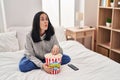 Hispanic woman eating popcorn watching a movie on the bed making fish face with mouth and squinting eyes, crazy and comical