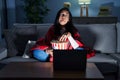 Hispanic woman eating popcorn watching a movie on the sofa making fish face with lips, crazy and comical gesture