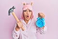 Hispanic woman with pink hair wearing pajama holding hammer and alarm clock afraid and shocked with surprise and amazed Royalty Free Stock Photo