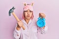 Hispanic woman with pink hair wearing pajama holding hammer and alarm clock puffing cheeks with funny face Royalty Free Stock Photo