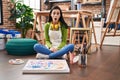 Hispanic woman sitting at art studio painting on canvas scared and amazed with open mouth for surprise, disbelief face Royalty Free Stock Photo