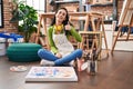 Hispanic woman sitting at art studio painting on canvas smiling happy and positive, thumb up doing excellent and approval sign Royalty Free Stock Photo