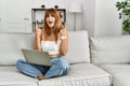 Hispanic woman sitting on the sofa at home using laptop amazed and surprised looking up and pointing with fingers and raised arms Royalty Free Stock Photo