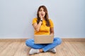Hispanic woman using laptop sitting on the floor at home afraid and shocked, surprise and amazed expression with hands on face Royalty Free Stock Photo