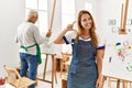 Hispanic woman wearing apron at art studio smiling and confident gesturing with hand doing small size sign with fingers looking Royalty Free Stock Photo