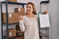 Hispanic woman working at small business ecommerce holding clipboard with blank space pointing thumb up to the side smiling happy Royalty Free Stock Photo